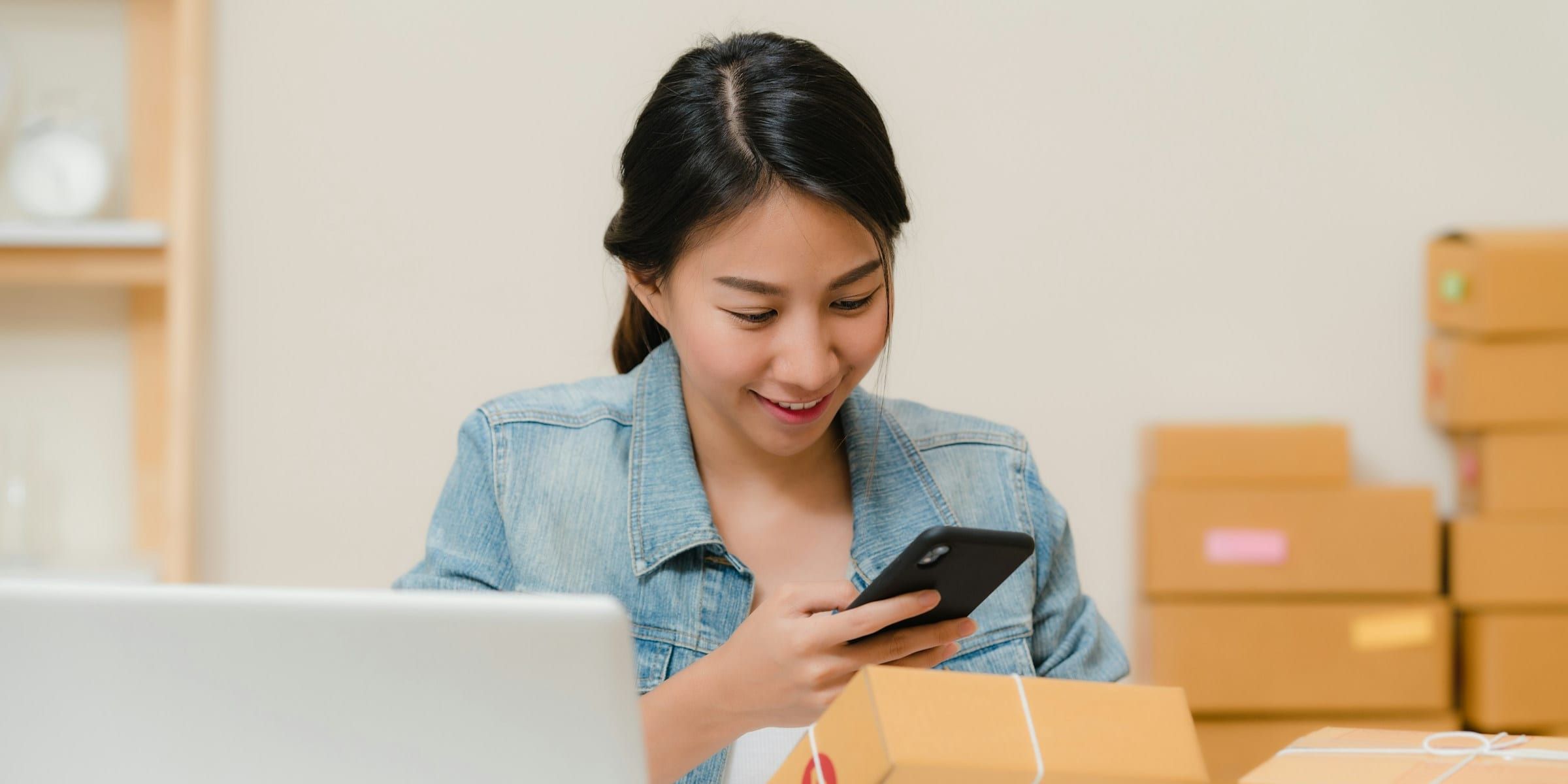 Small business owner at a desk scanning a packed parcel with her phone, a laptop beside her and order boxes stacked behind