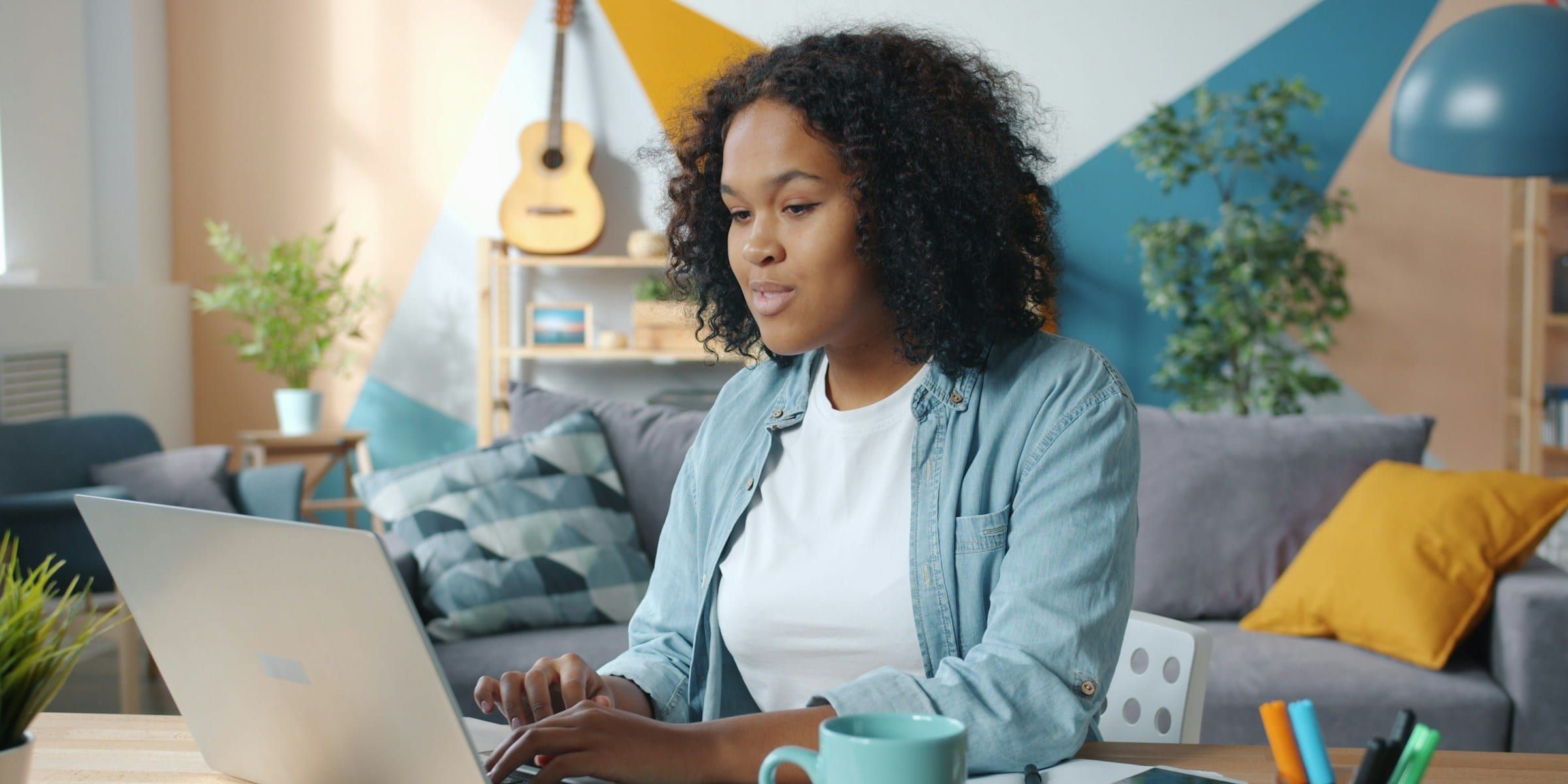 Woman typing on a laptop at a wooden table in a bright living room, with a mug, a notebook and a phone beside her